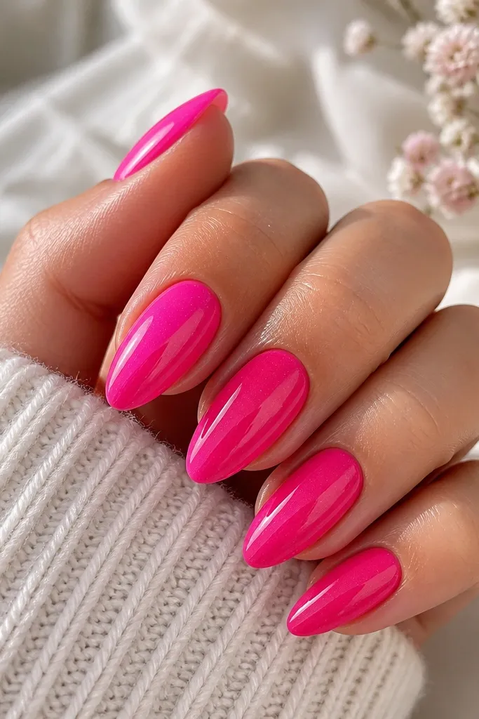 A set of almond nails painted a solid, high-gloss hot pink. The top coat has fine, nearly invisible micro-glitter that catches light as a soft sparkle, with crisp cuticle edges and a clean, reflective shine.