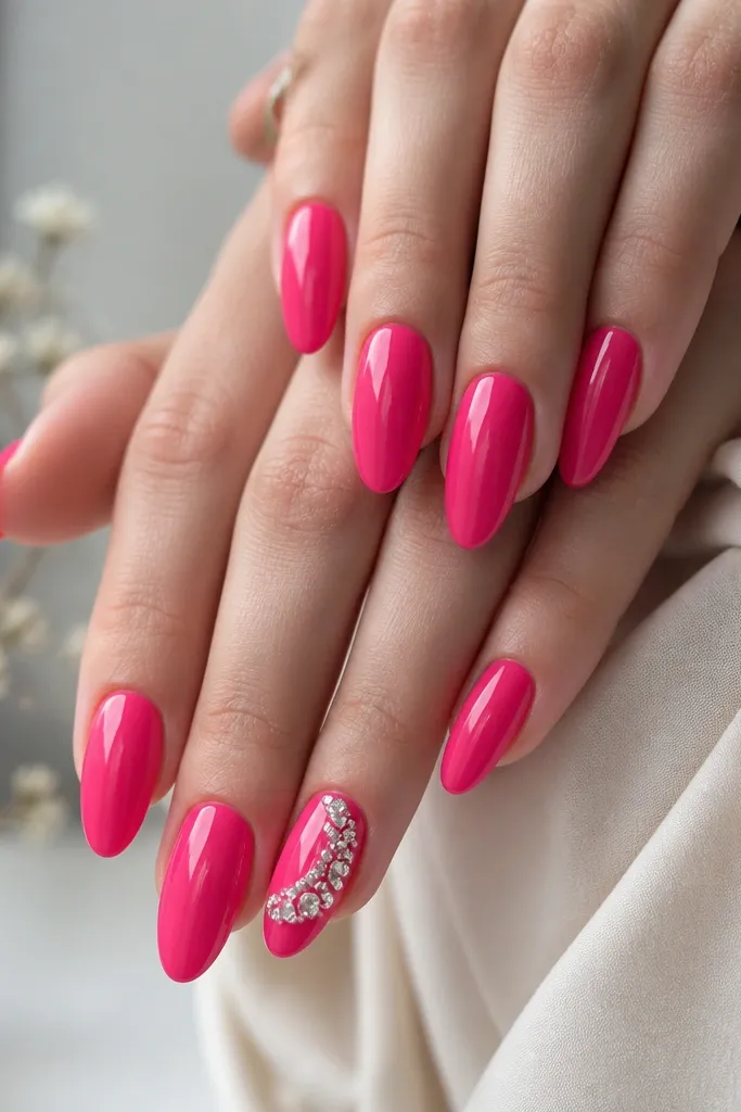 Hot pink glossy almond nails with one accent nail featuring a cluster of clear rhinestones on the sidewall near the middle and lower part of the nail. The rhinestones catch light, and the rest of the nails stay solid hot pink.