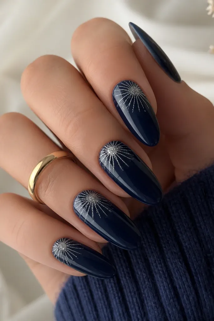 Long oval nails in deep midnight blue. The tips have a starburst pattern made from fine silver glitter radiating from the center outward, like small fireworks. The rest of the nail is glossy and smooth.
