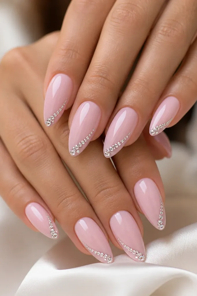 Almond nails in a soft powder pink base with a single diagonal track of tiny clear crystals along one side of each nail. The crystals start near the middle and angle toward the tip, with a few stones clustered at the apex area. Everything is glossy with crisp cuticle edges.