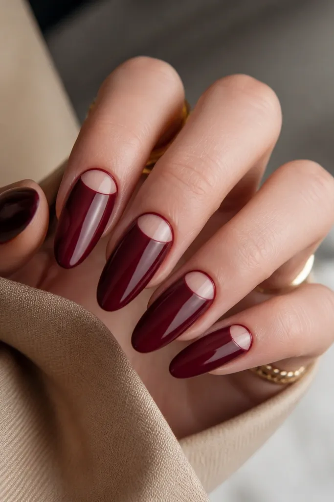 Almond nails with a glossy burgundy base. At the cuticle, there's a negative space half-moon where the nude nail shows through, outlined by a crisp burgundy edge. The rest of the nail is fully burgundy, with clean symmetry and sharp lines.