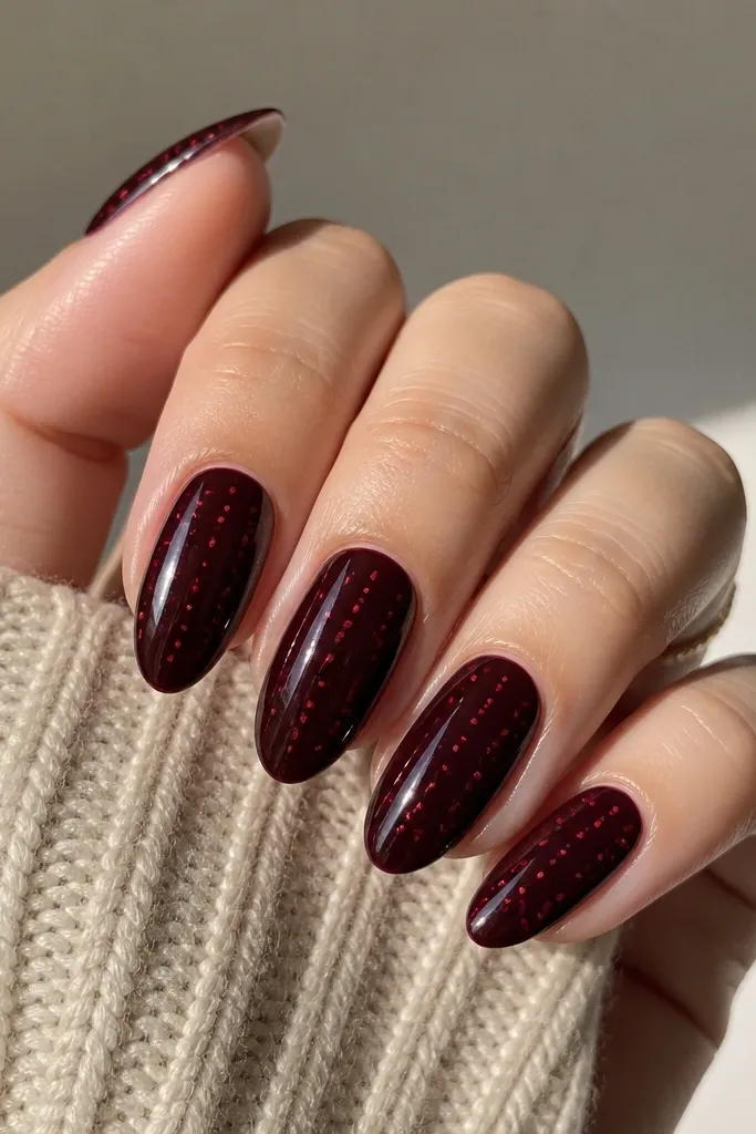 Almond nails in a glossy black cherry red base with scattered tiny red dots near the center and toward the tip. The dots are small, evenly spaced, and look like fine confetti. Each nail has a slightly different dot placement but stays balanced. Glossy finish.