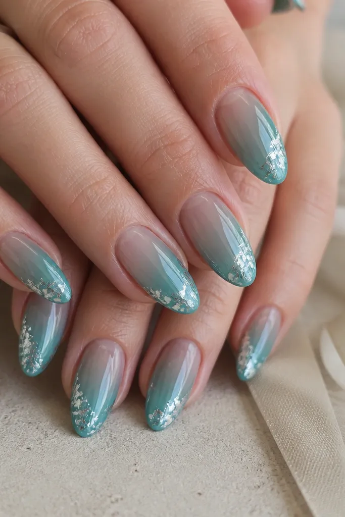 Almond nails in a translucent teal "glass" finish with silver foil flakes placed only at the outer corners near the tip. The foil looks like small torn pieces catching light at the edges. The center stays clear and glossy, and the foil doesn't cross into the middle.