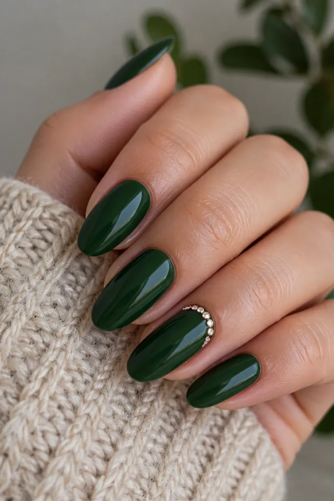 Almond nails in forest green BIAB with a glossy finish. On the ring finger and maybe one accent, there is a small crescent of tiny rhinestones near the cuticle, arranged in an arc. The stones catch light without covering the whole nail.