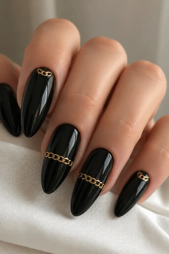 Long almond nails in glossy black. Two nails show a gold chain link pattern across the center, with tiny connected oval links. Other nails have a single gold link at the tip or near the cuticle.