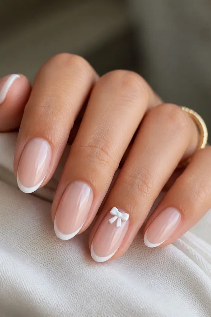 Short almond nails with a sheer nude BIAB base and classic white micro French tips. The ring finger has a tiny 3D bow in white near the cuticle, with the rest of the nails plain and glossy.