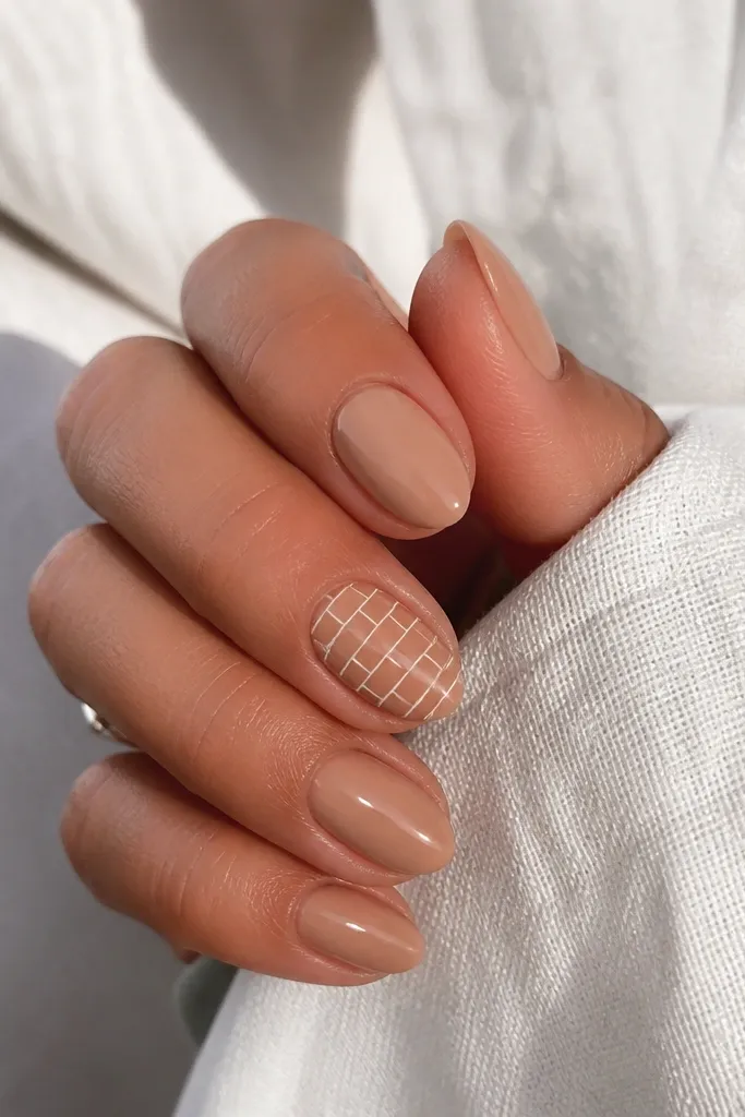 Short almond nails in warm tan nude BIAB. The ring finger has white brick-like lines forming small rectangles, like a tiny wall pattern. Other nails are plain tan.