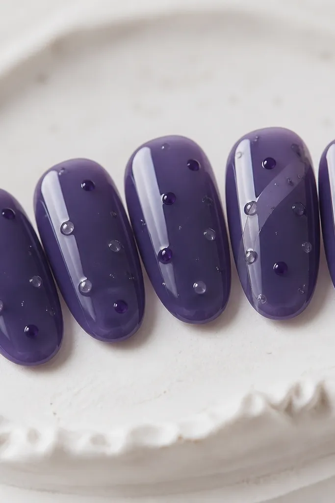 Five nails with a deep blueberry purple base that looks glossy and slightly translucent, like a glaze. Over it, there are tiny clear gel dots and a few darker purple dots placed like berry seeds. One nail has a subtle diagonal lighter glaze stripe. All nails are high shine.