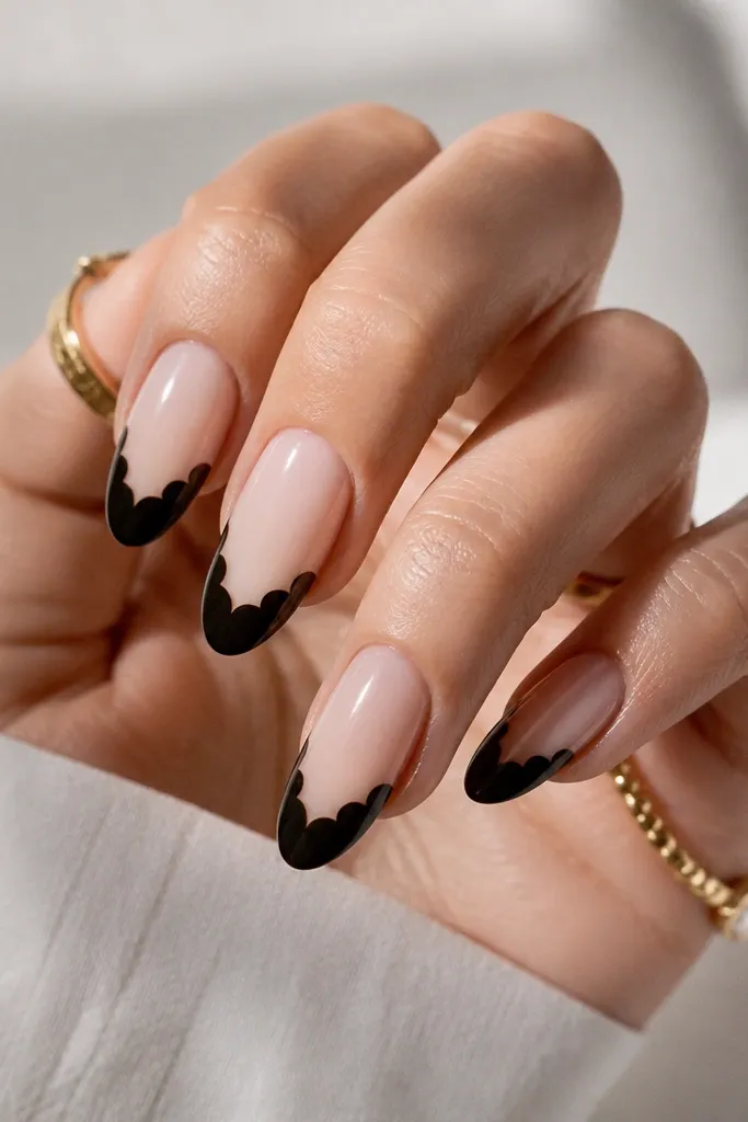 Almond nails with a sheer nude base and a glossy black scalloped tip. The scallops are evenly spaced like a fish scale pattern, and the black tip has a shiny, curved border.