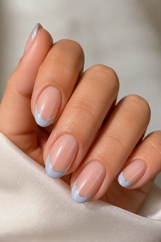 Short almond nails with a sheer nude base, a crisp icy blue French tip, and a thin line of fine silver micro-glitter only at the outer edge of the blue.