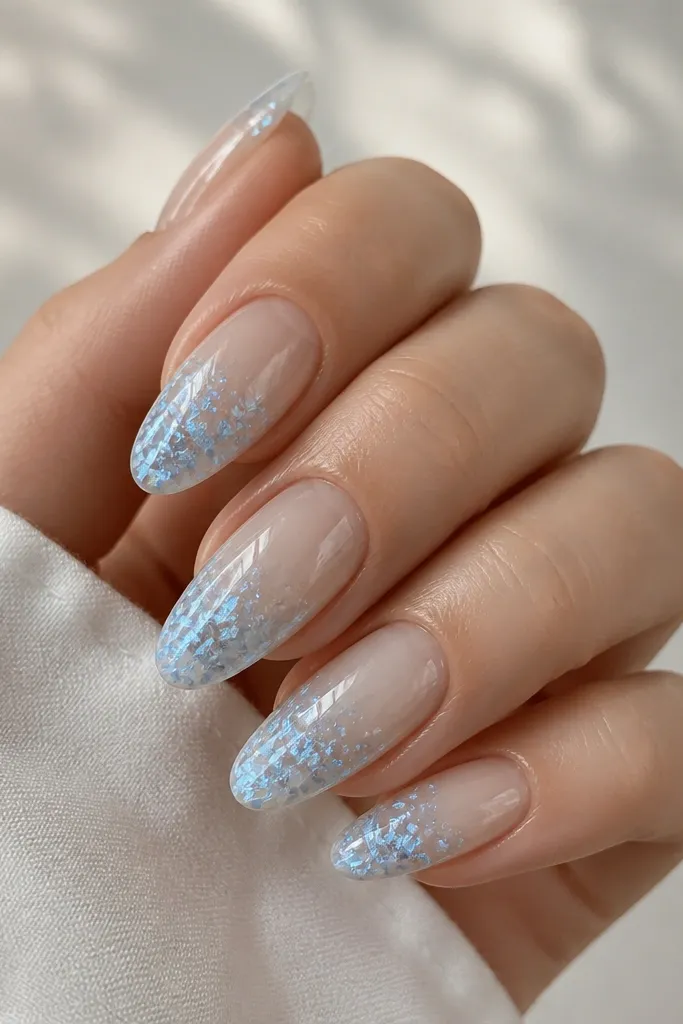 Medium almond nails with a clear or milky base and glacier blue foil flakes clustered toward the tip; the rest of the nail is mostly transparent.