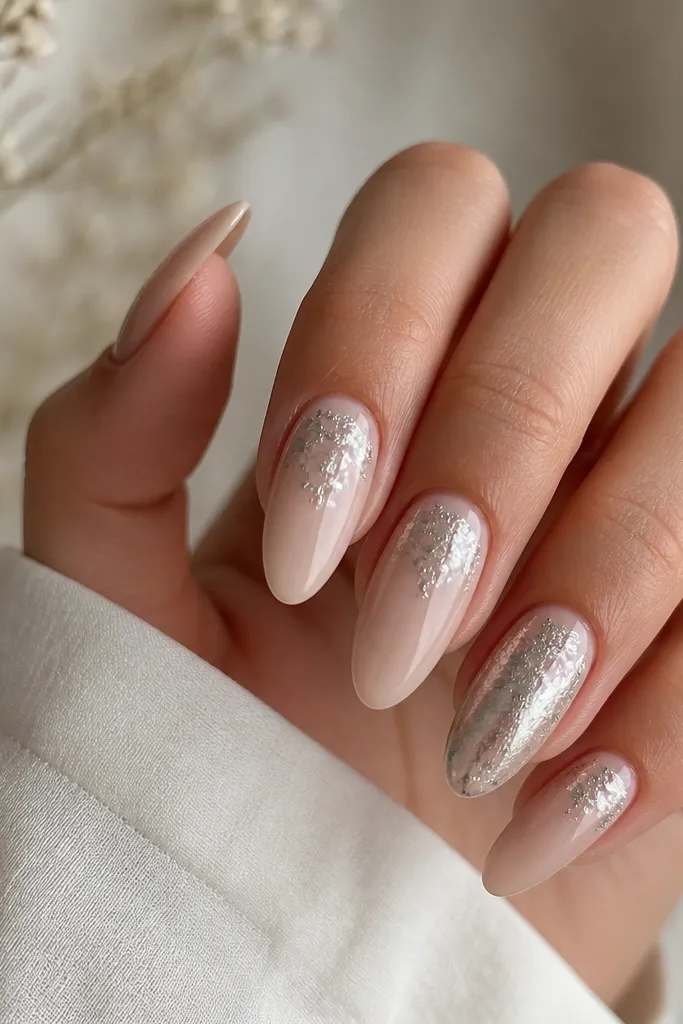 Almond extensions with a milky nude base. Over the nails, there are wispy thin streaks of silver foil arranged like a veil across the top third of each nail. The edges of the foil look feathered, not solid blocks. One nail has slightly heavier foil placement.