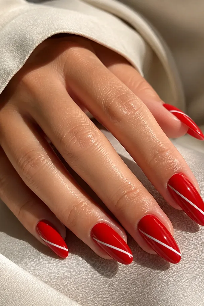 Almond nails in bright red with one or two thin white stripes running diagonally from near the sidewall toward the center. The stripes are crisp and evenly spaced, with glossy finish.
