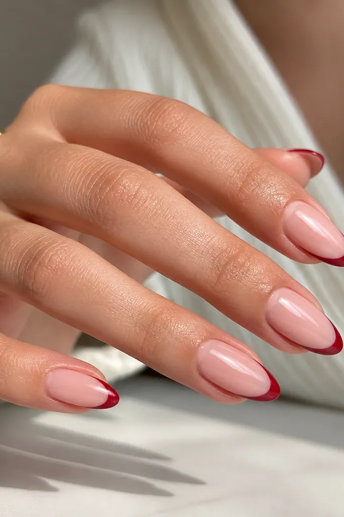 Almond nails with a sheer nude base and a thin cherry red French tip; the smile line is crisp and slightly curved, with glossy topcoat.