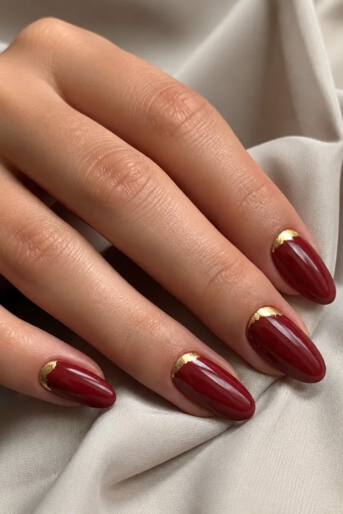 Cherry red almond nails with small irregular gold foil crescents near the side of the cuticle; glossy finish.