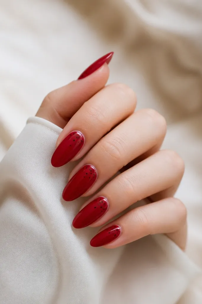 Glossy cherry red almond nails with scattered tiny black dots like a small constellation near the upper third.