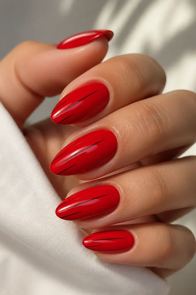 Glossy red almond nails with one or two very thin black lines per nail, lines are vertical and slightly angled, they look like fine pen strokes and stay near the center.