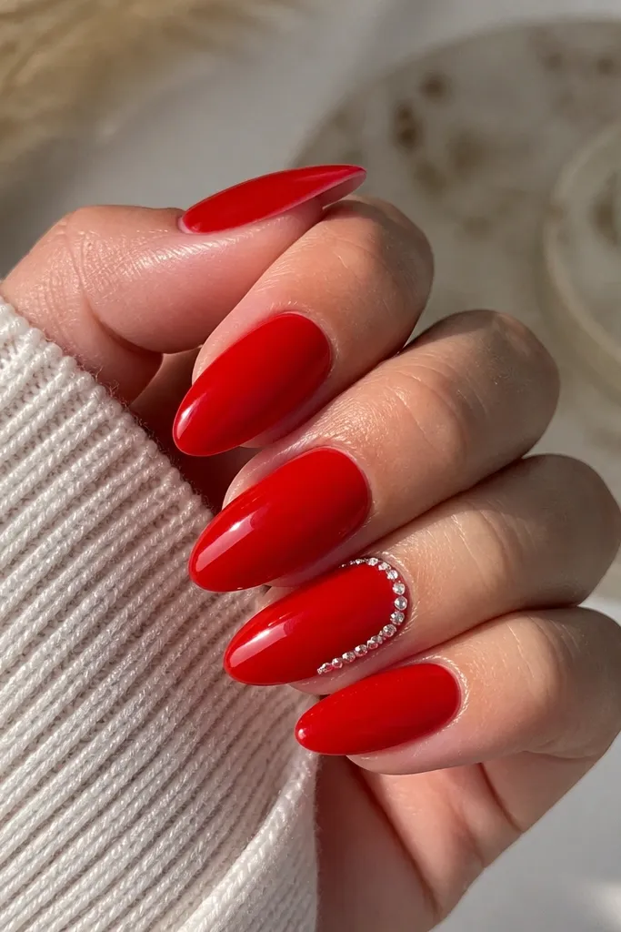 Most nails are glossy red almond. One accent nail has a row of small clear rhinestones along the side cuticle edge, forming a half-border curve, with clean spacing and a reflective sparkle.