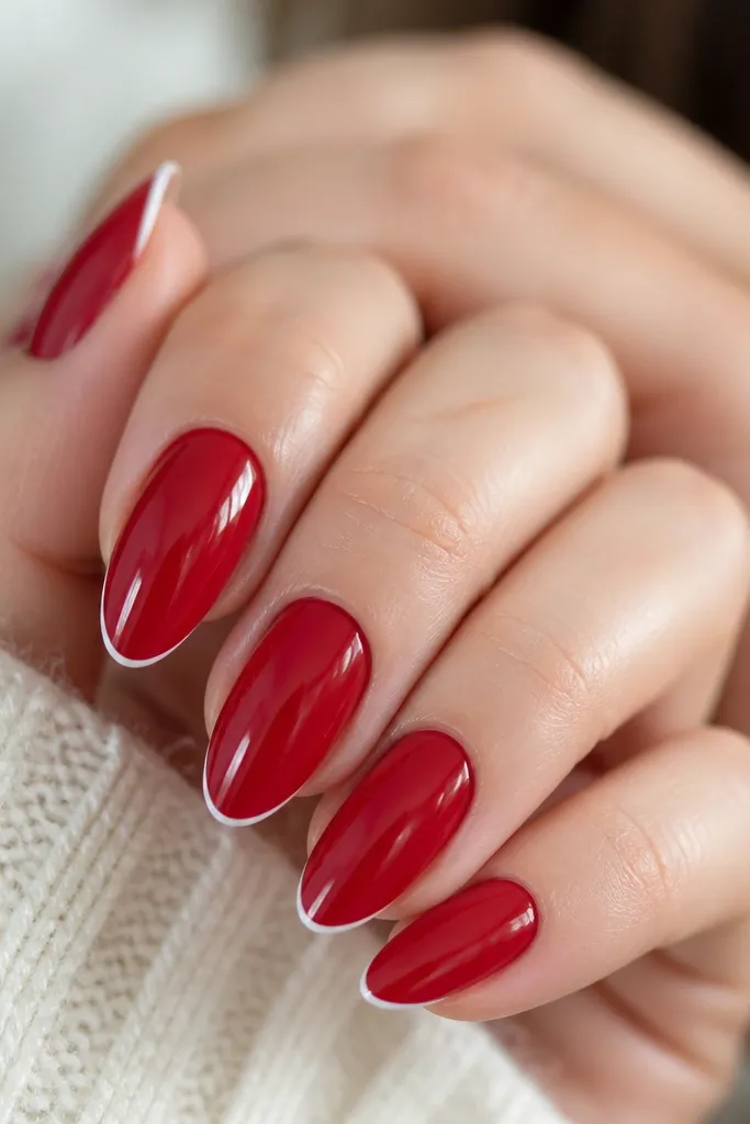 Close-up of almond nails painted cherry red with ultra-thin white French tips. The tips are narrow, following the almond curve, and the cuticle area stays clean with a glossy finish.