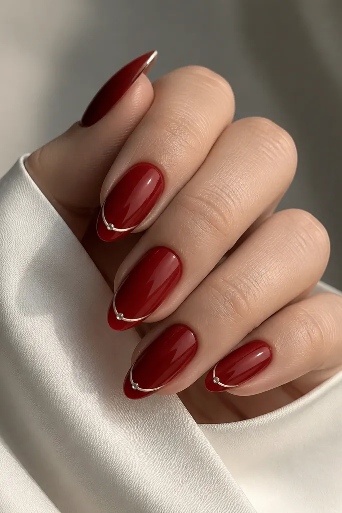 Almond nails with deep red base and a thin French line. Each nail has a single small rhinestone placed at the outer corner of the French line, all on a glossy background.