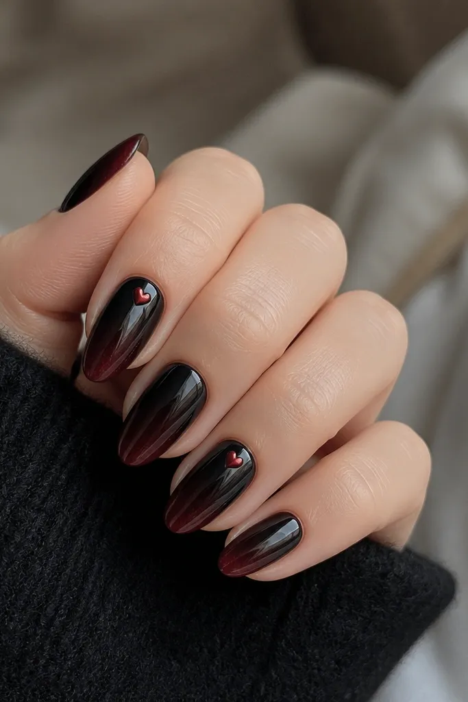 Short almond nails painted black at the base fading into a warm cherry tint at the tips. Two nails have tiny red heart shapes punched with a dotting tool, placed near the cuticle. The rest has a glossy finish with subtle red shimmer.