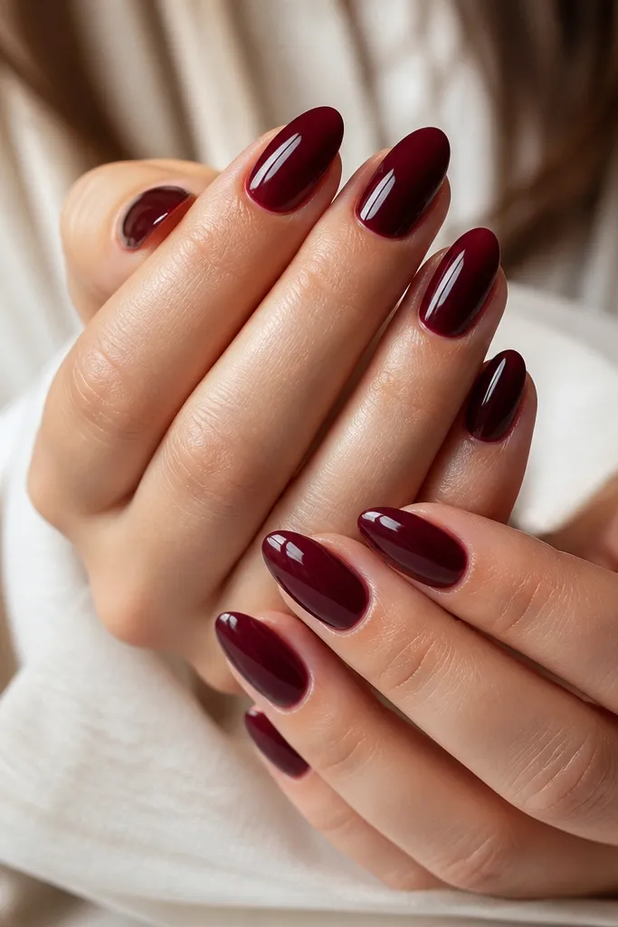 A full set of almond nails painted a deep cool burgundy with a mirror-like shine. The nails are medium length and the cuticle area looks crisp and clean, with smooth, even color from base to tip.