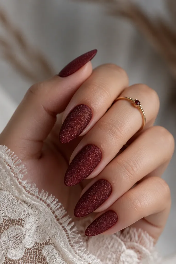 Almond nails in a dark red with a soft, velvety matte texture. The nails look like they have tiny crushed-grain sparkle, with no mirror shine and a smooth, even surface.