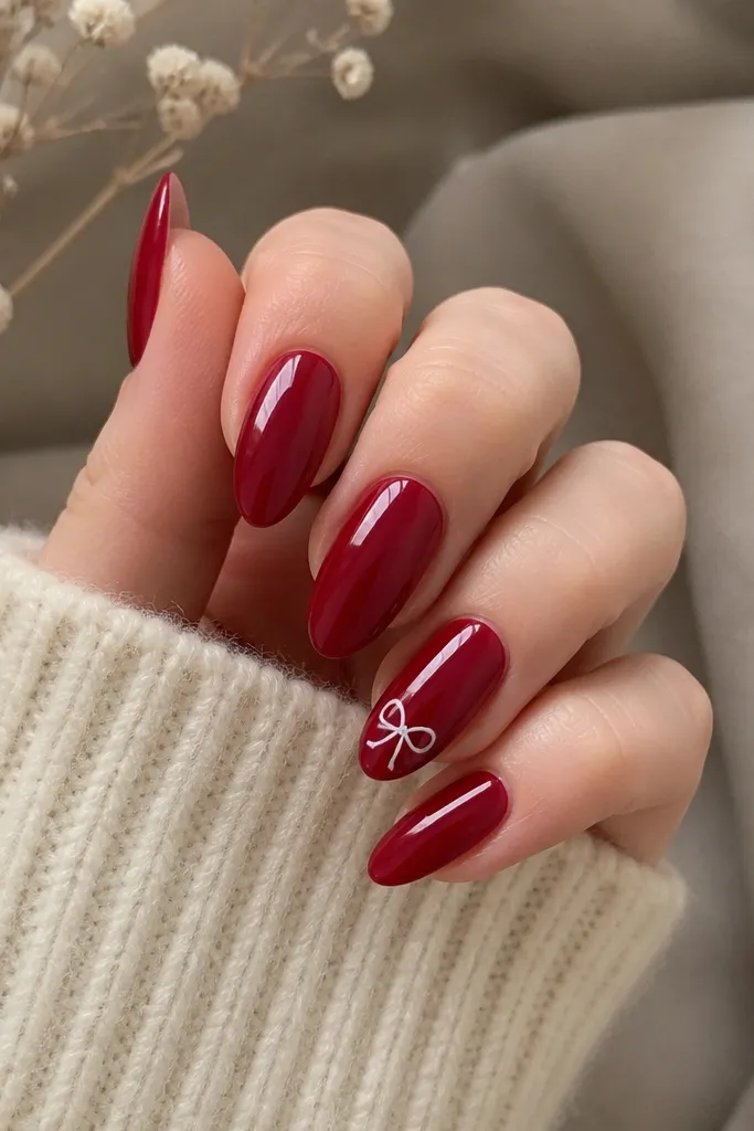 Almond nails in solid dark red with one accent nail showing a small white bow outline near the tip. The rest of the nails are glossy and plain, so the bow stands out.