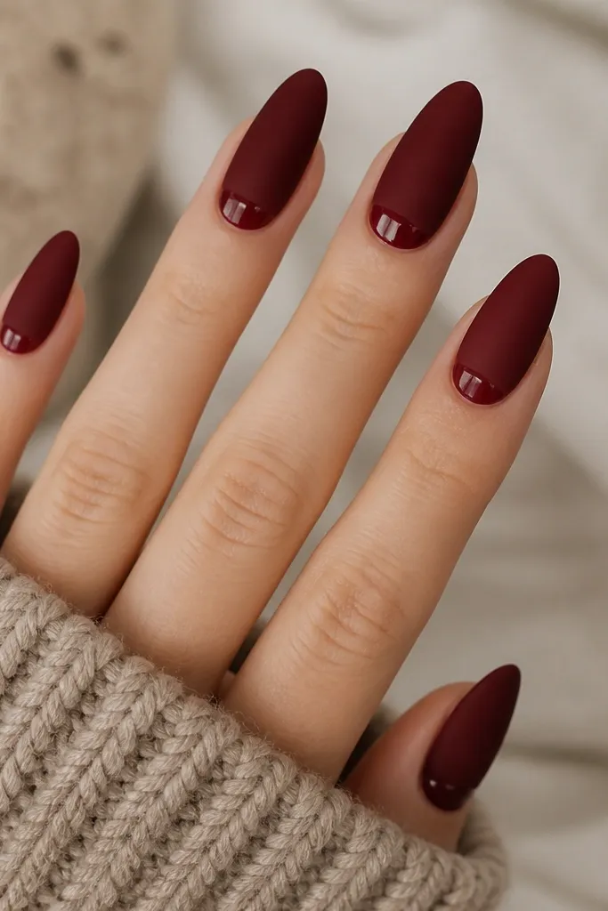Almond nails painted dark red matte. A glossy dark-red half-moon is visible at the cuticle area on each nail, creating a shiny crescent contrast.