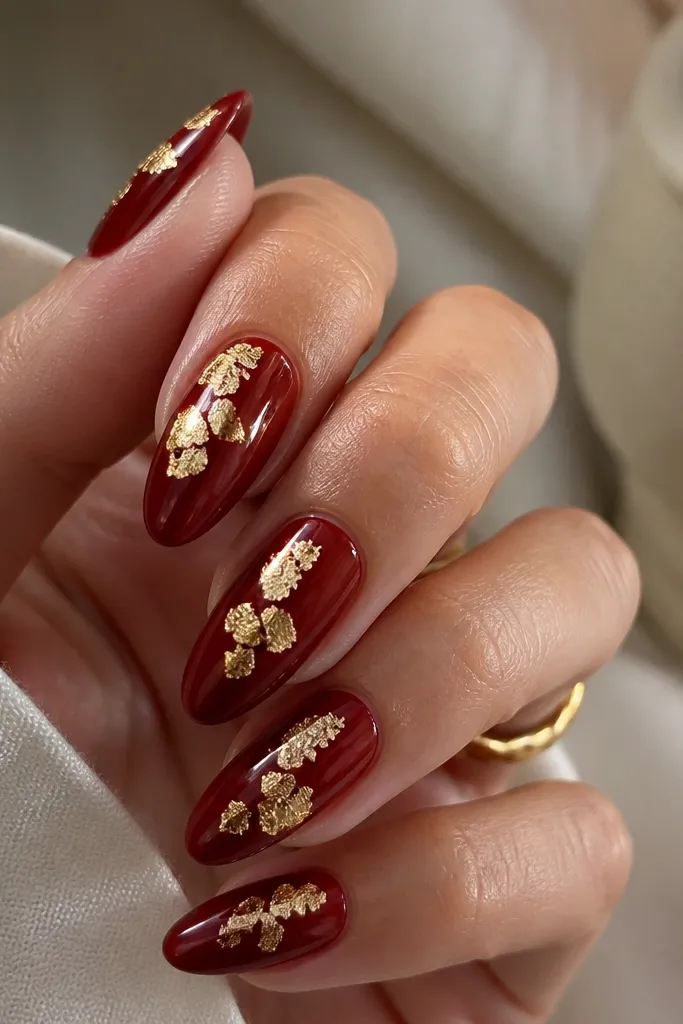 Almond nails painted dark red with irregular gold leaf pieces placed near the center and slightly toward the sides. The gold looks matte-gold with a glossy red background underneath.