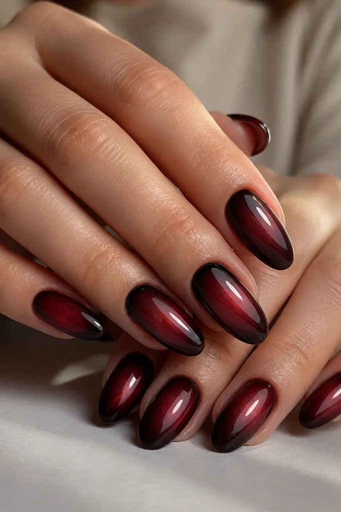 Almond nails in deep cabernet red with a smoky, airbrushed gradient that fades into near-black at the cuticle edges. The center glows with a soft red halo like a backlit wine stain, finished in a high-gloss top coat. One accent nail has a thin, curved black arc tracing the cuticle line.