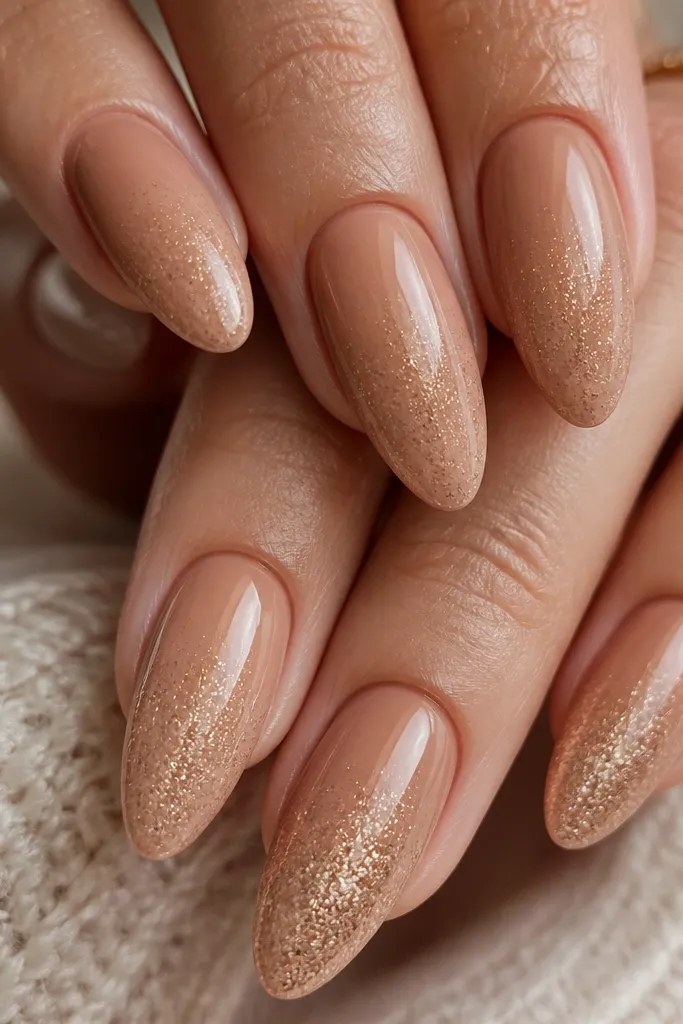 Close-up of almond nails painted a creamy cinnamon nude, with a soft honey micro-glitter fade starting around mid-nail and tapering to the tips. Glossy top coat reflects light; two ring nails have slightly heavier glitter coverage.
