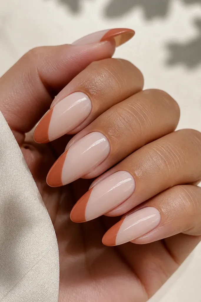 Almond nails with a sheer milky base. The tips show a terracotta French twist that curves diagonally, creating a modern asymmetry. Everything is ultra-glossy and clean around the cuticle.