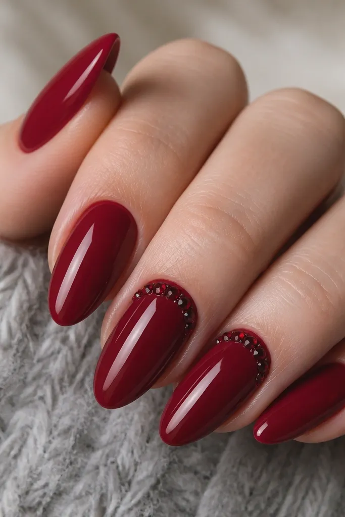 Almond nails painted autumn berry red with a glossy finish. On two nails, tiny blackened red crystals cluster near the cuticle in a small arc. Crystals catch light and create texture.