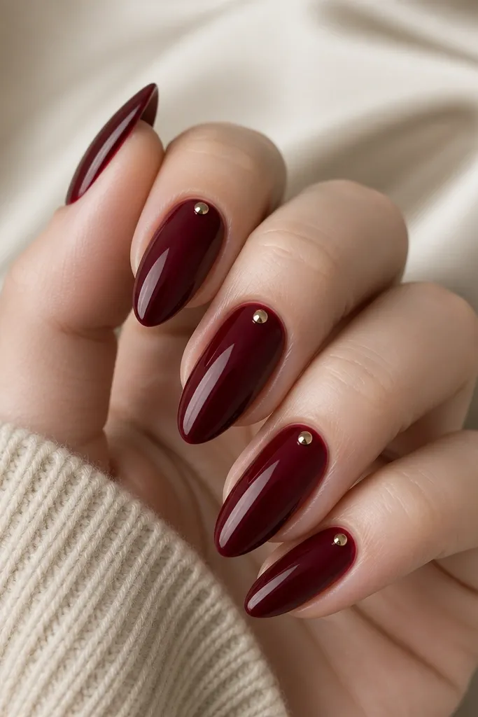 Almond nails in classic burgundy glossy lacquer. Each nail has one tiny rose-gold stud placed near the cuticle on the outer edge, like a single accent jewel. Clean negative space around the stud.