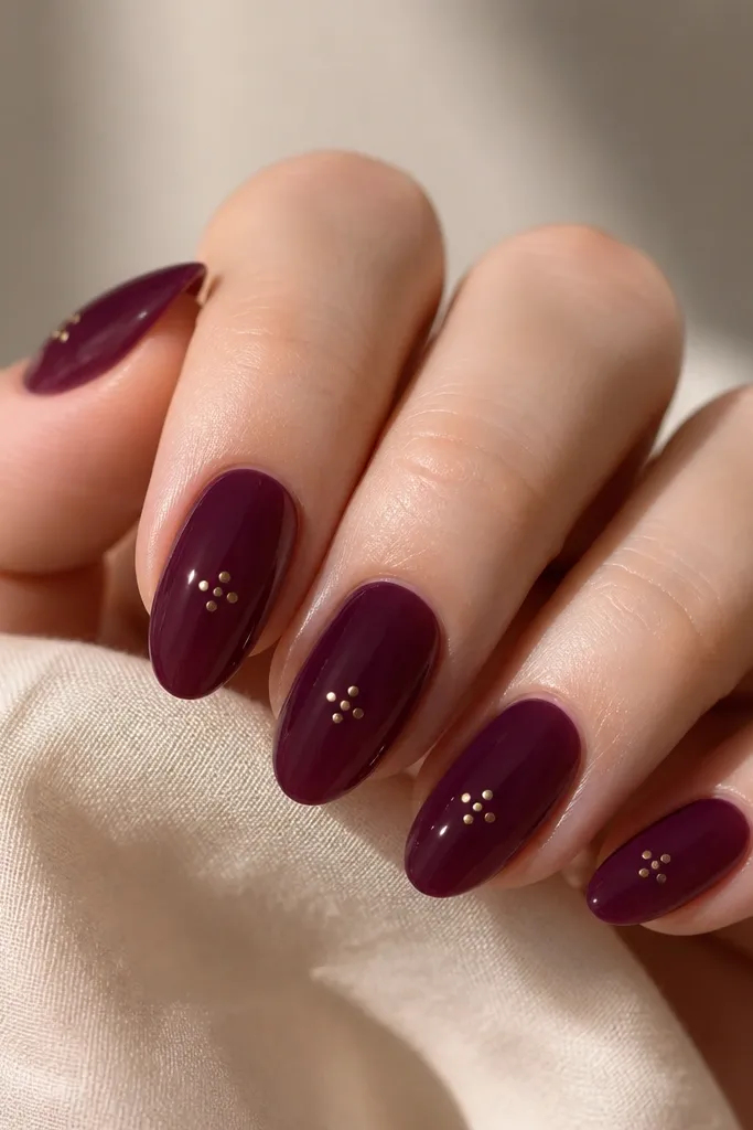 Almond nails in a deep plum jelly polish. On each nail, three tiny gold dots form a small triangle near the center. Glossy, glassy finish with smooth leveling.