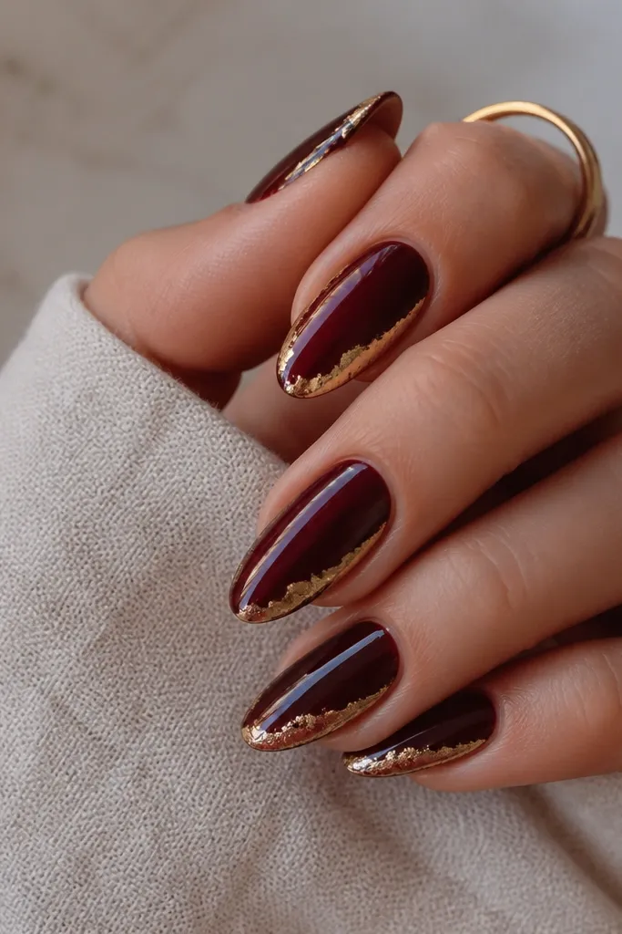 Almond nails painted deep oxblood glossy. Along the outer edge of each nail, a thin strip of irregular gold foil wraps from sidewall to near the tip. The foil looks like torn metal with natural edges.