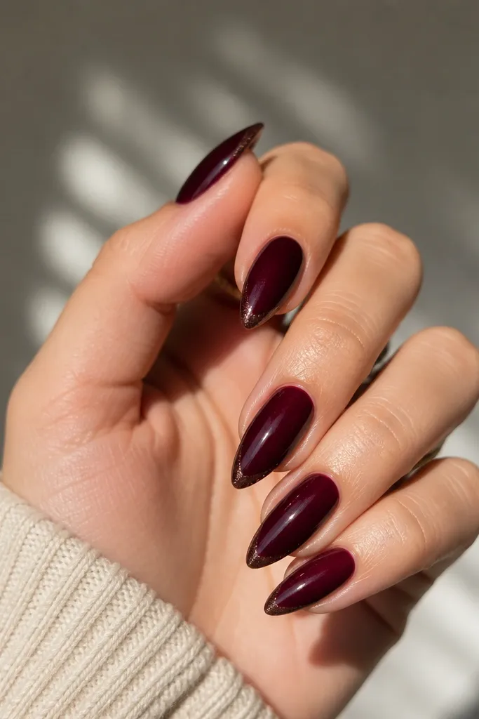Short nails painted deep oxblood. Each nail has a sharp V-shaped tip near the free edge, outlined by a thin line of warm micro-glitter, with a glossy finish.