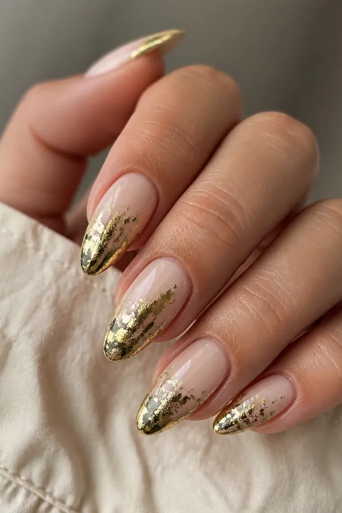 Long almond nails with a champagne nude base. The tips are covered in irregular green foil pieces that look like torn metal. The foil sits mostly on the tip area and blends slightly into the base, with a glossy finish.