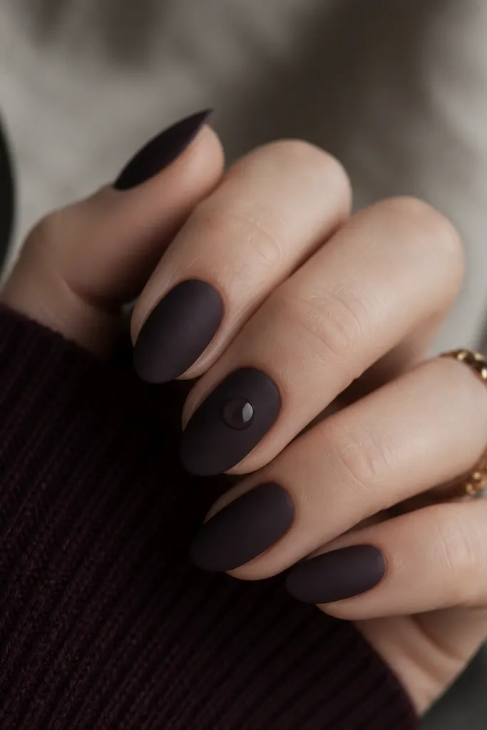 Short almond nails in blackened plum with a velvet matte finish; one nail has a single glossy clear dot near the center, while the others stay matte.
