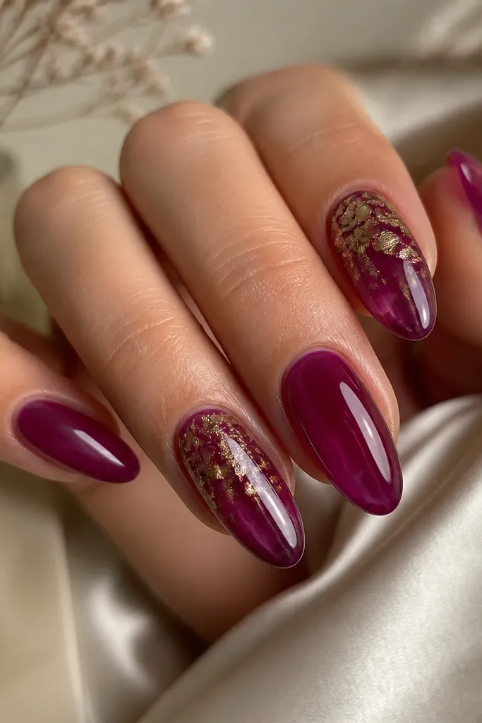 Medium almond nails with a translucent plum jelly base. Two nails have sheer gold foil applied to the upper half, like broken flakes. The foil is not fully opaque and sits over the plum for a stained-glass look.