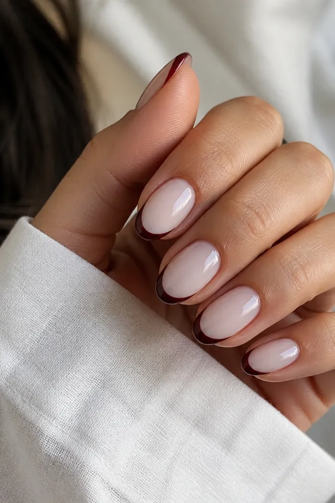 Short almond nails with a sheer milky nude base. The French tips are very narrow and painted oxblood red-brown, with a crisp curved line and glossy top coat. No glitter, just clean color blocking.