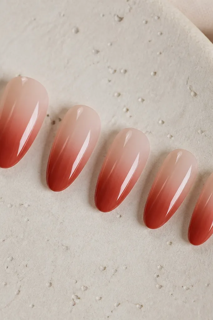 Five press-on nails in soft almond shape. The nails show a press-on ombre from sheer nude at the cuticle to brick red at the tip. The surface is glossy and smooth with no extra art.