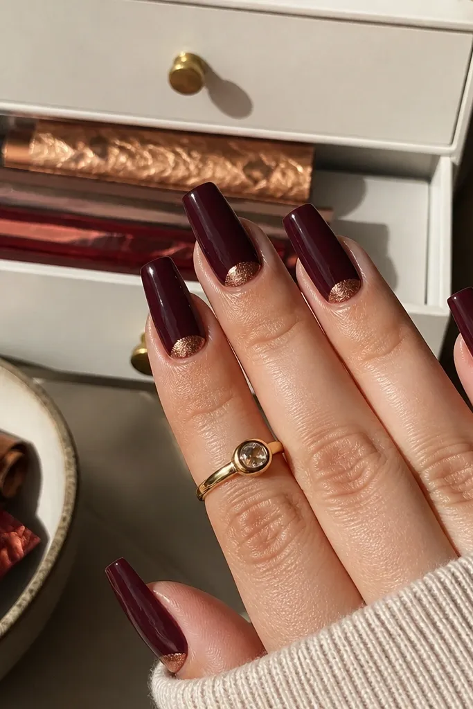 Coffin nails with deep oxblood dip. At the cuticle, a clean half-moon of copper foil sits like a crescent. The background shows a small drawer with foil sheets rolled and stored in a flat folder.