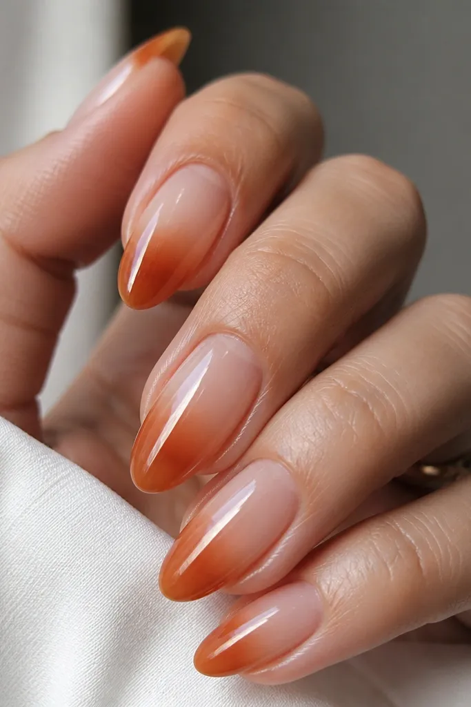 Medium-length almond nails with a clear jelly center. The tips fade from sheer nude into rust-orange, forming a smooth gradient. The nails look glossy and glassy, with the gradient concentrated at the outer half.