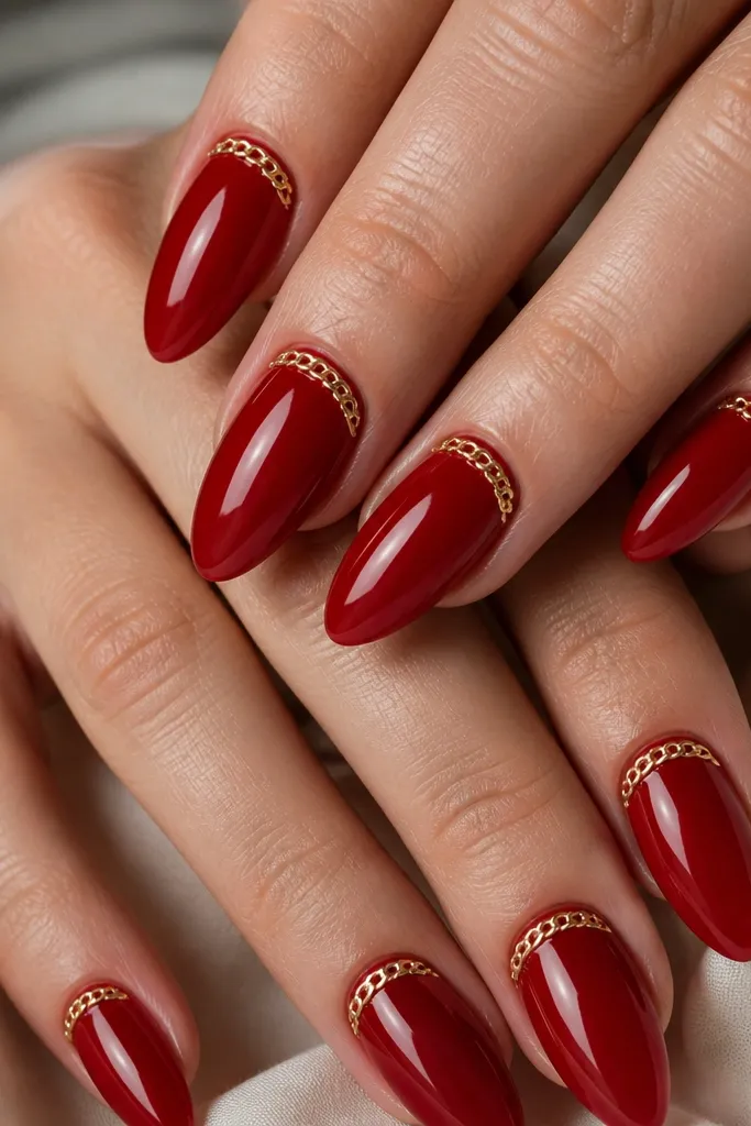 Long almond nails in a glossy classic red gel. Along the cuticle line, a thin gold chain detail wraps gently across the nail on most fingers. The gold chain is crisp and reflective, with a clean red border around it.