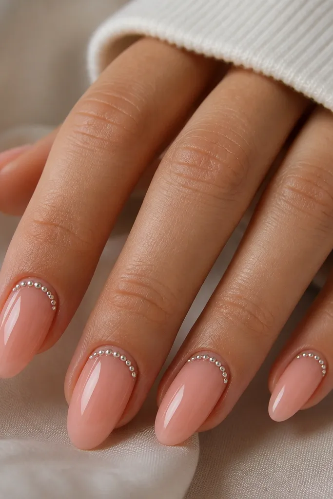 Medium almond nails in a peachy pink glossy base. A silver beaded half-moon border sits at the cuticle area, curving across the nail like a crescent. The beads are tiny and evenly spaced, with a clean peach edge around them.