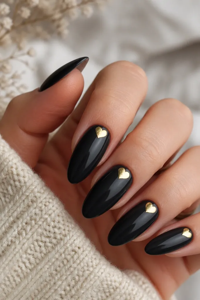 Long almond nails with a black milky opaque base. A small gold foil heart sits near the cuticle on each nail, with glossy finish.