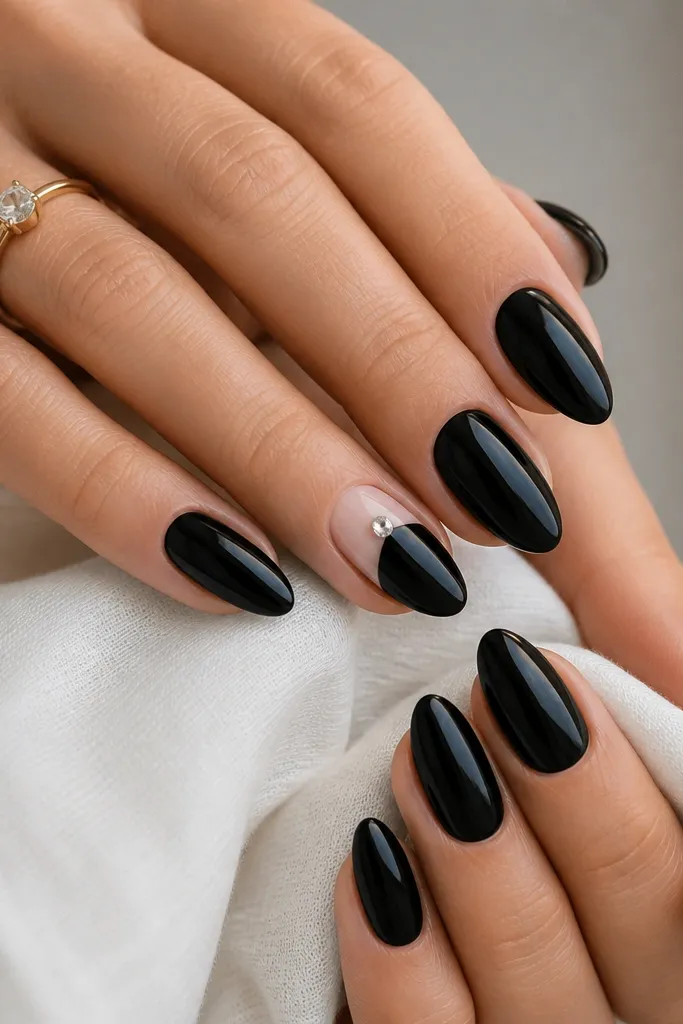 Glossy black nails in a medium almond shape. Each nail is plain except one accent nail with a single clear crystal placed near the cuticle on a sheer nude patch.