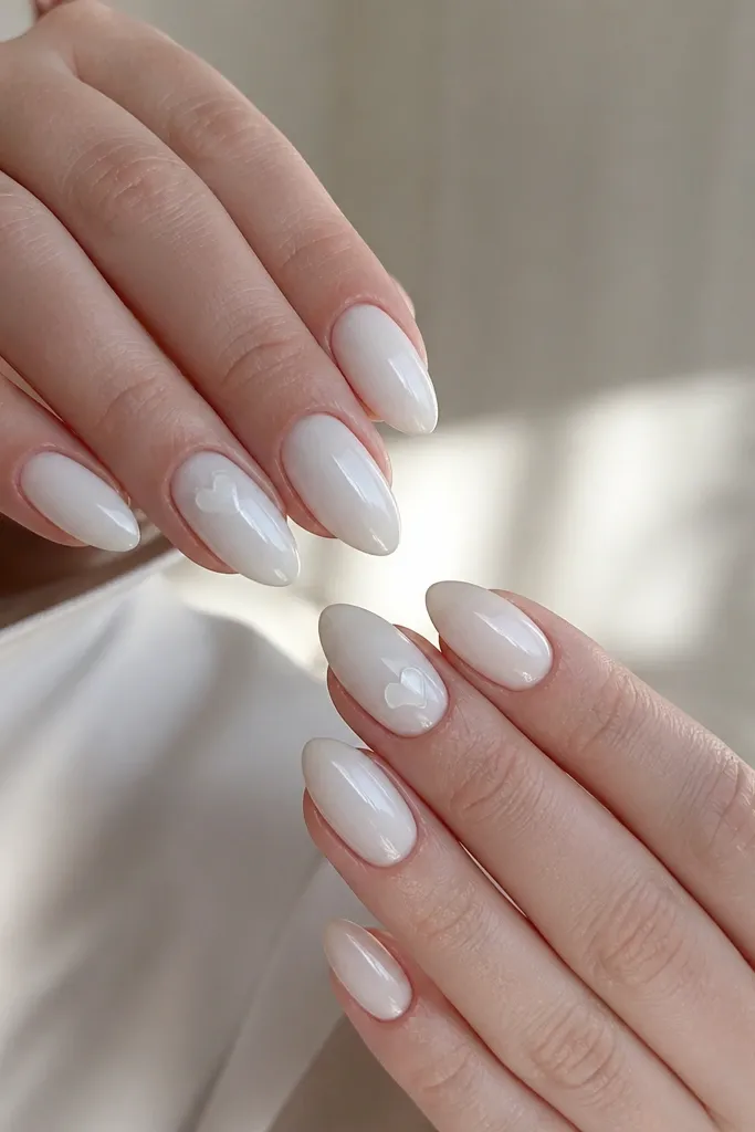 Almond nails with a milky white opaque base. On one nail per hand, a tiny white-on-milky or clear-on-white heart sits near the cuticle area, glossy and neat.